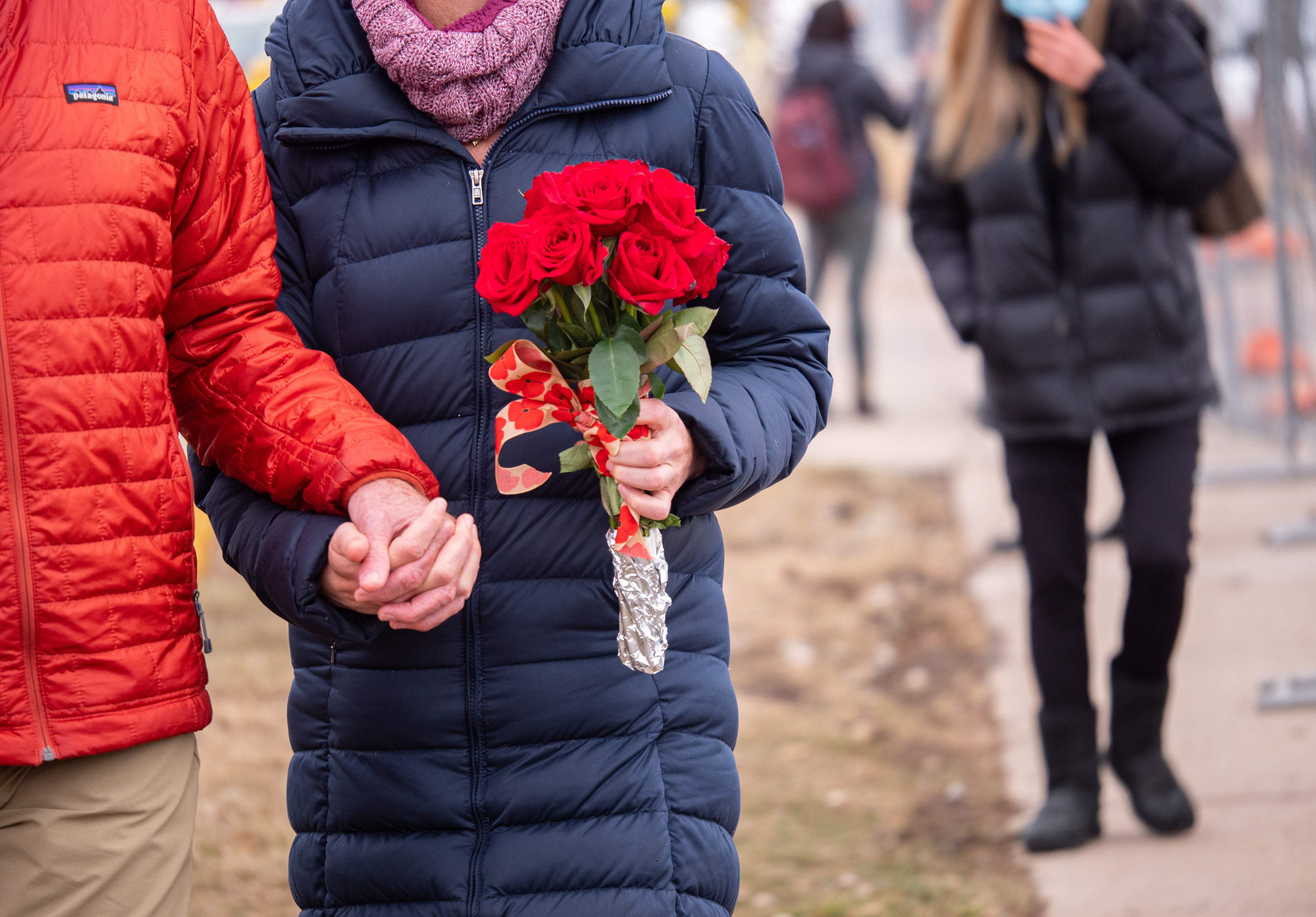 People hold hands outdoors near a sidewalk; one has a bouquet of flowers. 