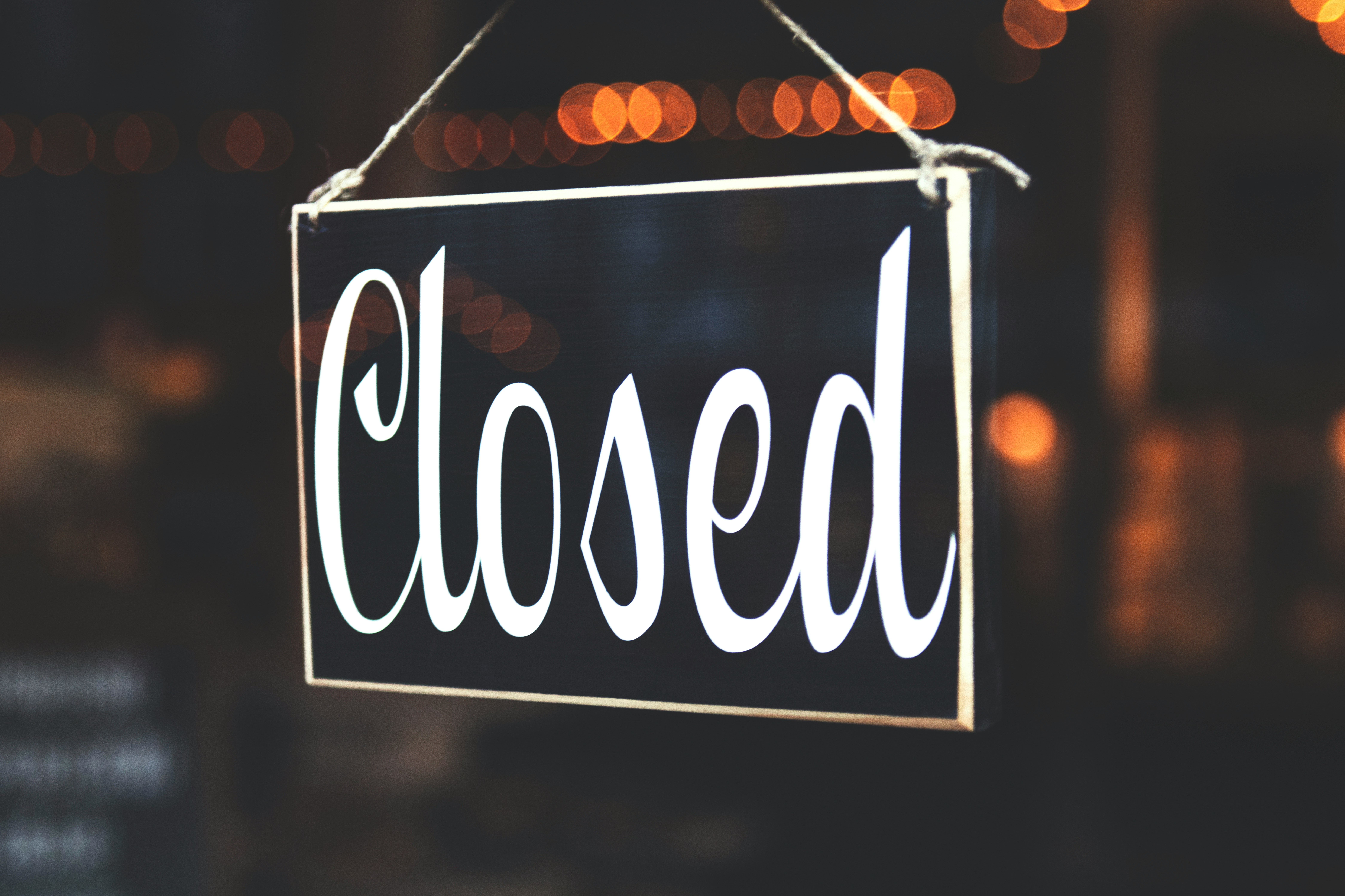 A black sign hangs in a window with the word "closed" in white and blurred orange lights in the background.